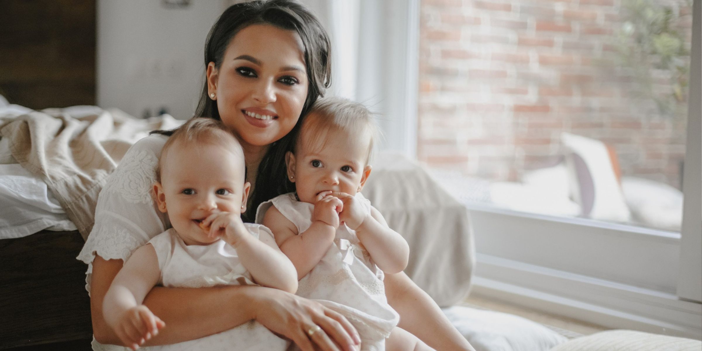 Mother smiling while holding two babies dressed in white outfits | Source: Magnific