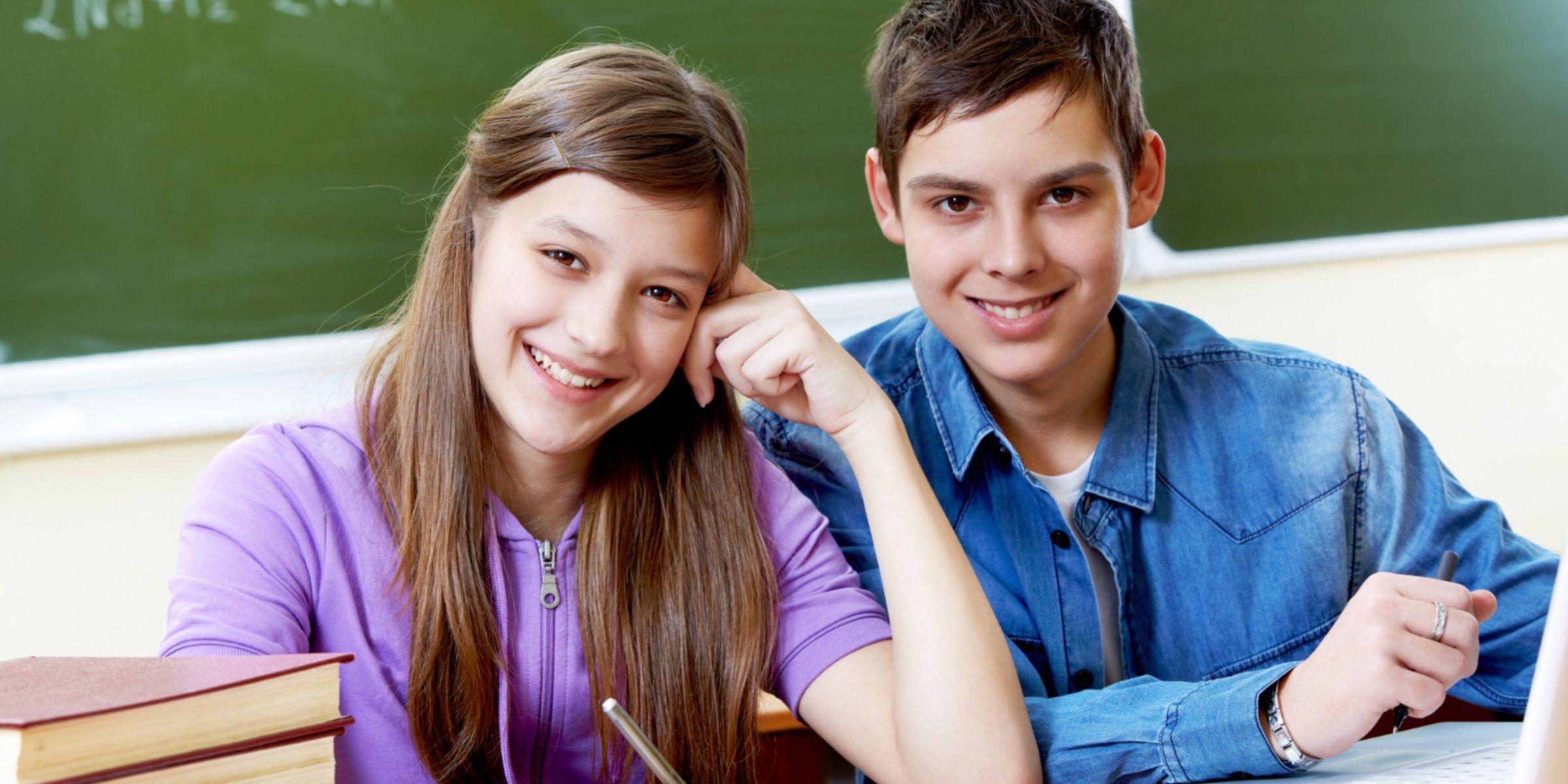 Two smiling students sitting together at a classroom desk with books and a laptop | Source: Magnific