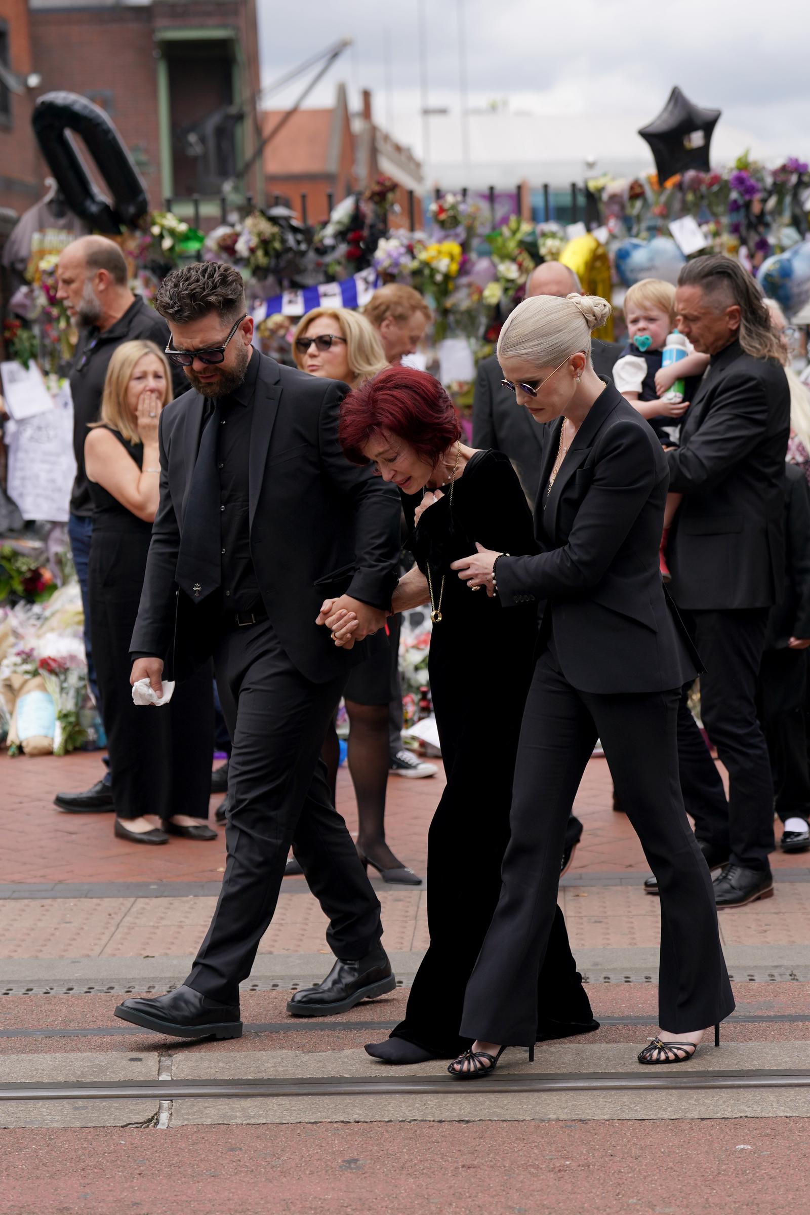 Jack, Sharon, and Kelly Osbourne view the messages and floral tributes left at the Black Sabbath Bridge bench on Broad Street in Birmingham, in memory of Black Sabbath frontman Ozzy Osbourne, as his body is brought back to his home city for a procession following his death at age 76. Picture date: Wednesday July 30, 2025. | Source: Getty Images